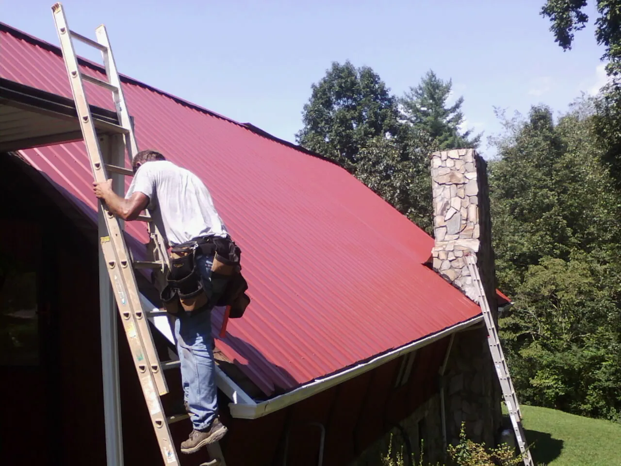 Roofer on a ladder beside a red metal roof and stone chimney — Soffit & Fascia Repair by Roofing Brothers in Carville